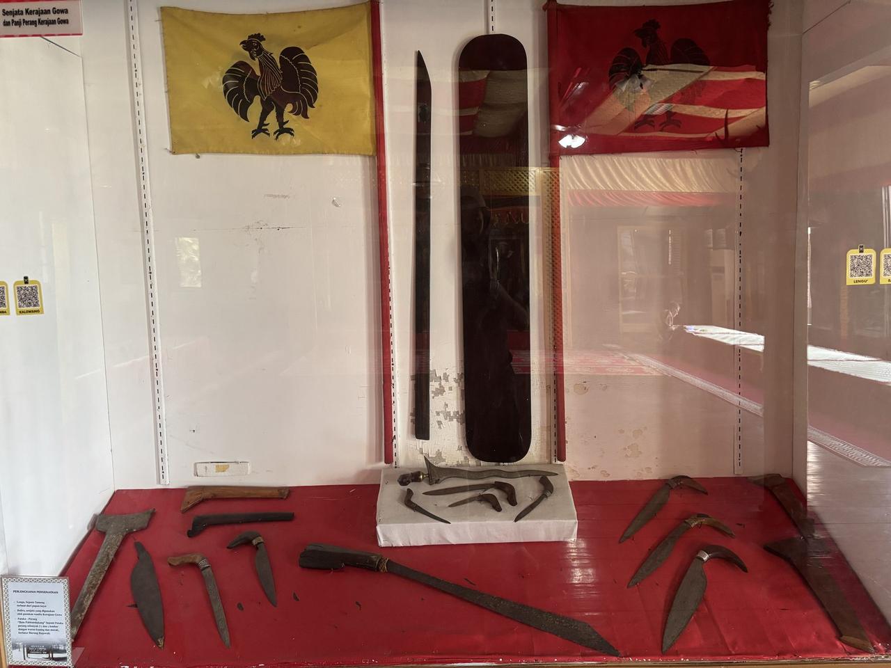 Bendera-bendera upacara dan sederetan senjata tradisional Gowa di dinding — bilah pedang, tombak, dan sebuah perisai anyaman — dipajang di balik kaca.
