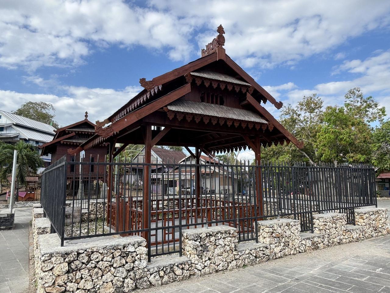 The wooden pavilion protecting Batu Popaua, built in traditional Buton architectural style with an ornately carved roof, fenced off within Benteng Keraton Buton.