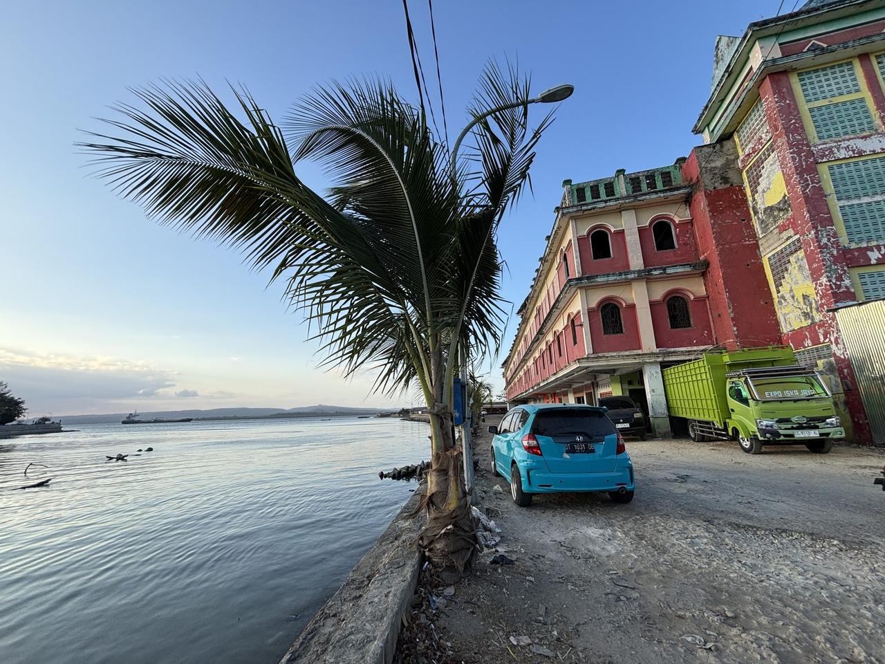 A weathered red and cream colonial-style building standing at the water's edge in Baubau, a truck and car parked in front.