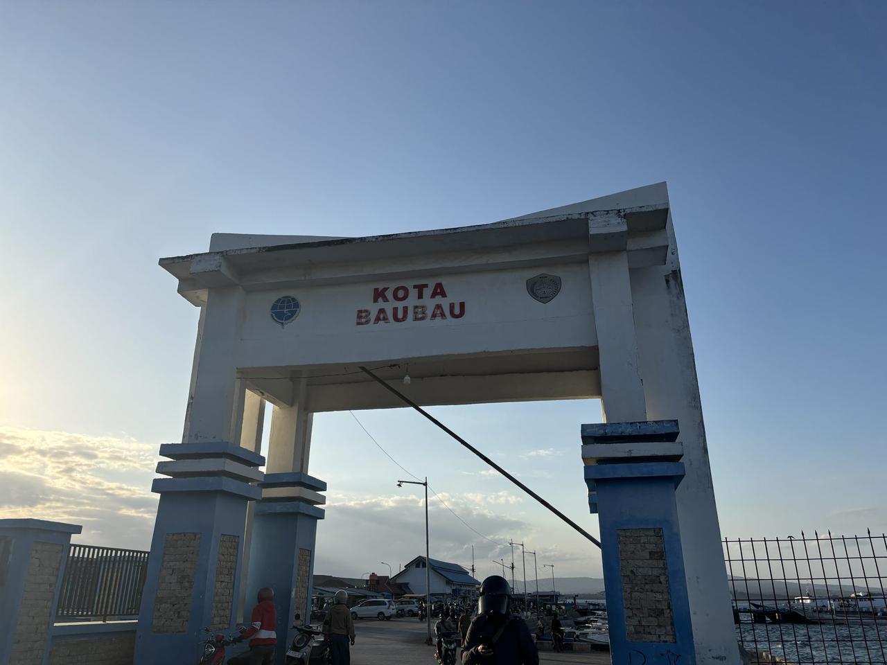 An arch reading Kota Baubau standing near the harbor, motorbikes passing beneath it in the evening light.