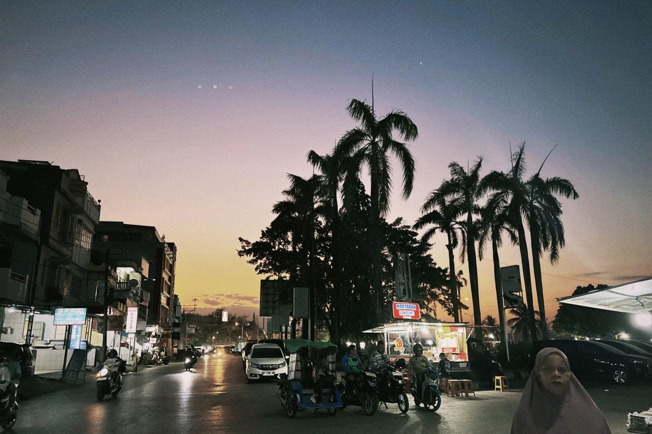 A street market coming alive at dusk in Baubau, food stalls and motorbikes beneath tall palm trees against an orange and blue sky.