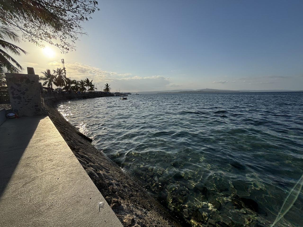 The waterfront at Baubau at sunset, clear water lapping against a rocky breakwater beneath palm trees.