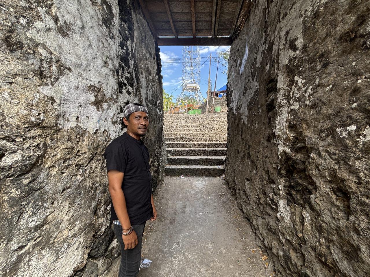 A guide standing in a narrow stone gateway inside Benteng Keraton Buton, with a lookout tower visible in the distance beyond a staircase.