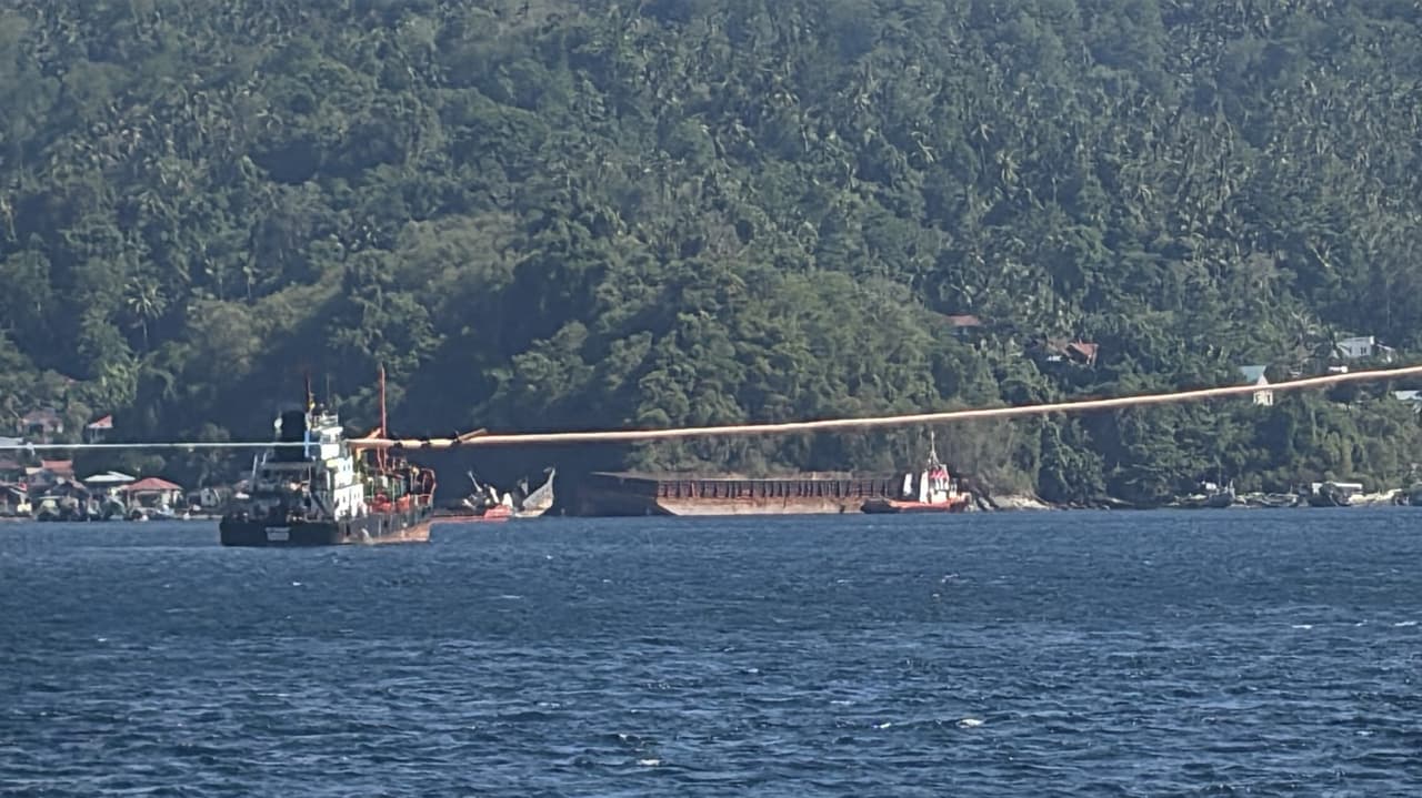 A boat being towed along the shoreline near Bitung's harbor, green hills in the background.