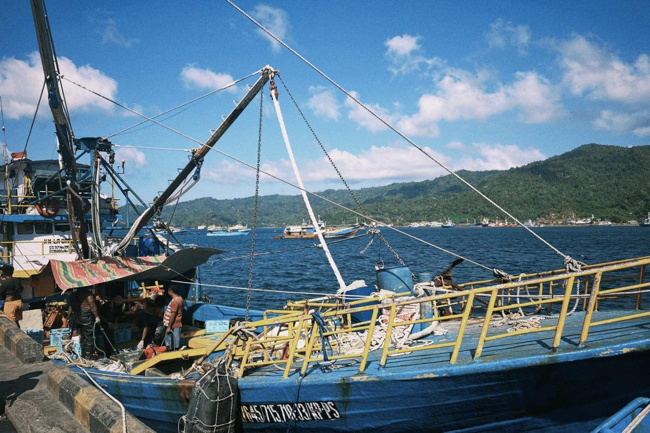 Crew members unloading fish from a boat onto a truck at the Bitung harbor, rigging and mountains visible behind them.