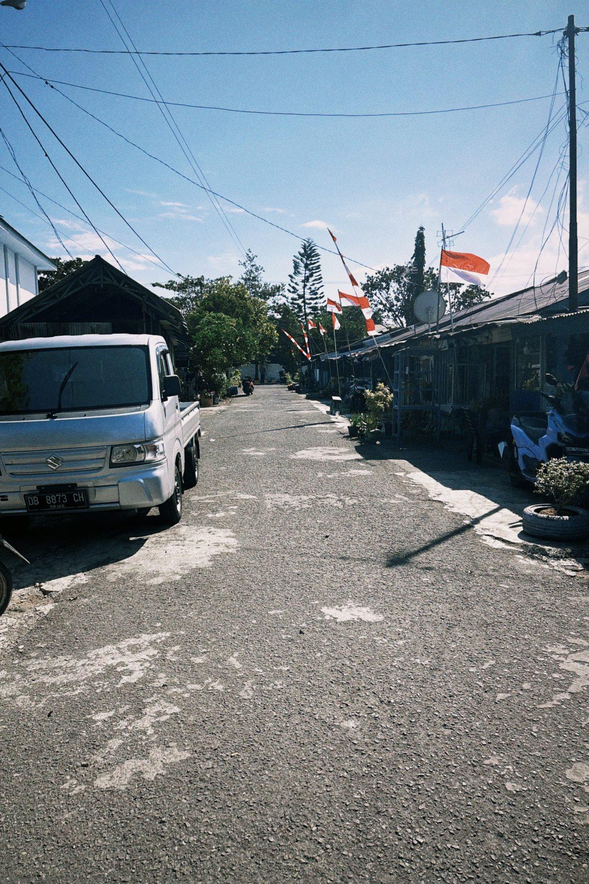A village street lined with red and white flags on the way to the Bitung fish market, a truck passing shopfronts.