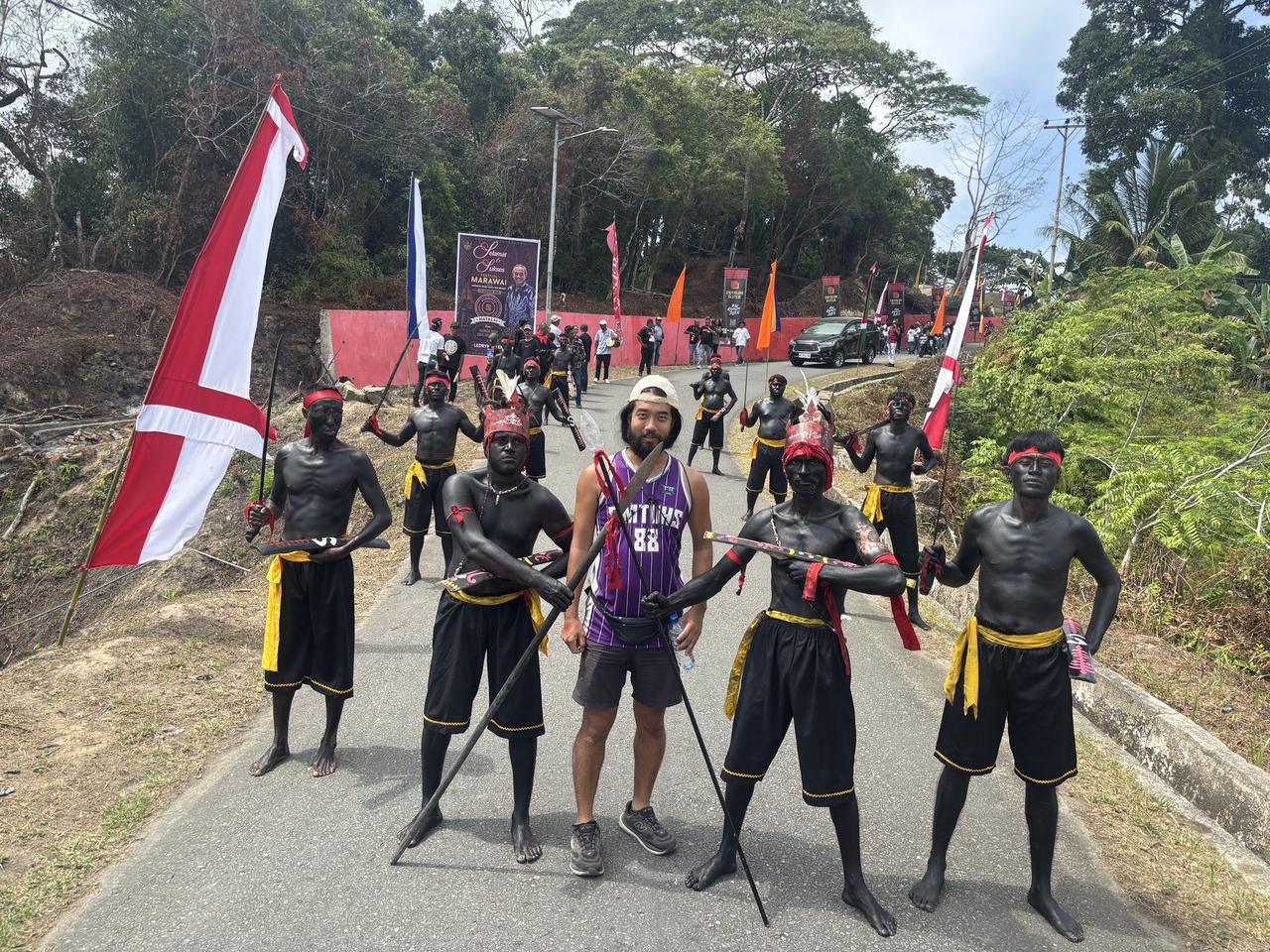 The author standing among Cakalele dancers with bodies painted black, holding real blades and Indonesian flags on the road outside Festival Marawai in Hatalai village.
