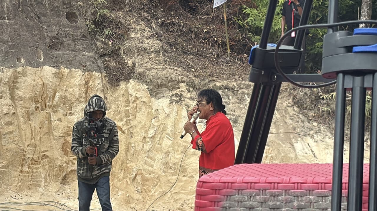 An older performer in a red shirt singing into a microphone on stage at Festival Marawai in the evening, with a cameraman filming beside him.
