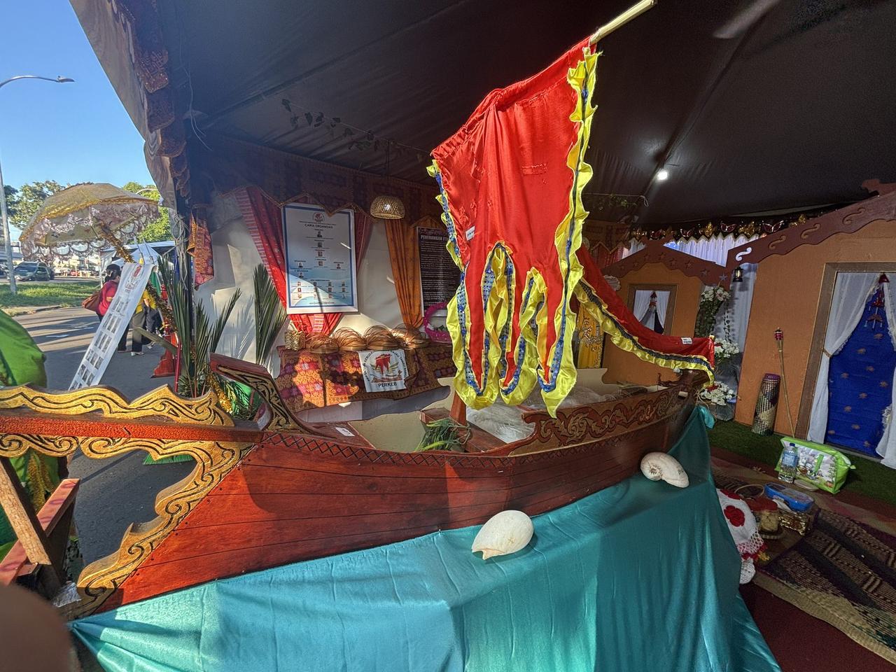 Inside a festival tent, a traditional wooden boat draped in teal cloth with a red and yellow flag mounted above it, seashells resting along its edge.