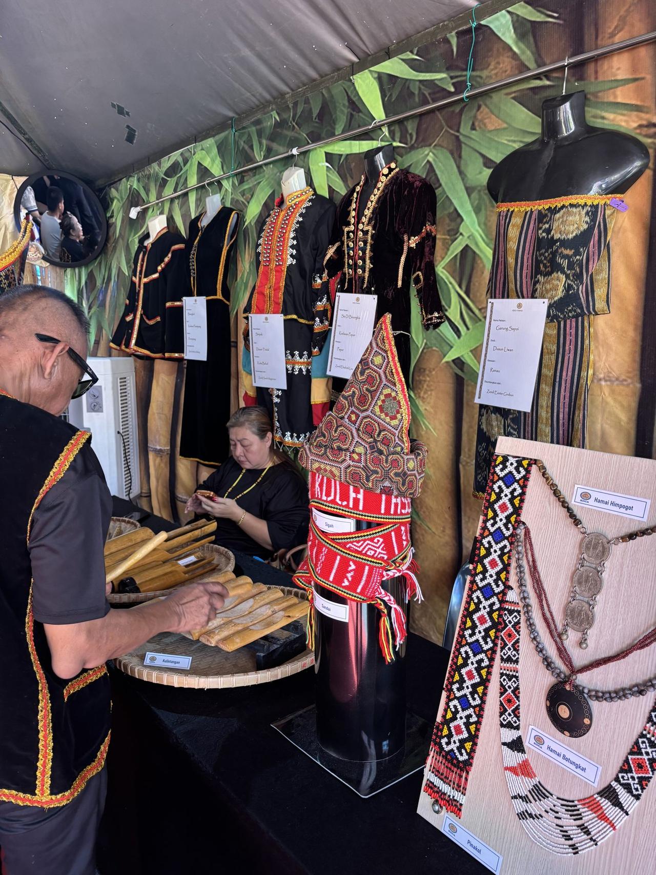 A Kadazan Dusun cultural booth with traditional costumes hung by district, a man arranging a row of kulintangan gongs, and beaded jewelry displayed on a board.