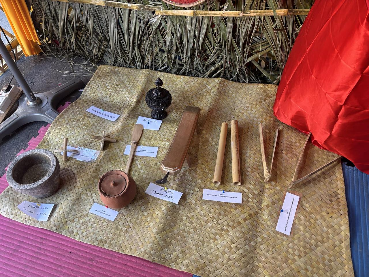 A woven mat display of traditional Kaili kitchen tools, each item labeled by name, including a stone mortar, a water container, and a coconut grater.