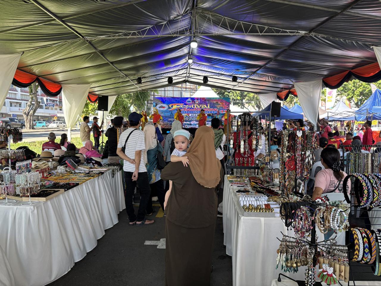 A covered market aisle inside the festival grounds, stalls of jewelry and accessories on either side, a screen showing the festival's name in the background.