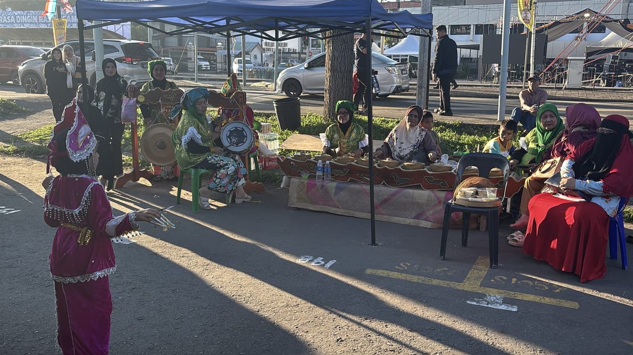 A group of women in traditional dress playing gongs and a drum at a roadside setup, facing a young dancer in a pink costume performing in front of them.