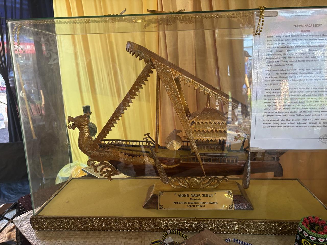 A gilded model dragon boat called Ajong Naga Sekui displayed in a glass case, beside a historical synopsis panel about the ancient Tidung kingdom.