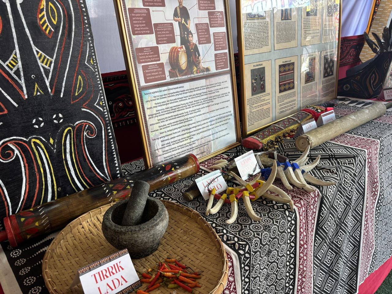 A Toraja display of a keris, a boar-tusk bracelet, a stone mortar and pestle, and a small woven basket labeled Tirrik Lada holding red and green chilies.