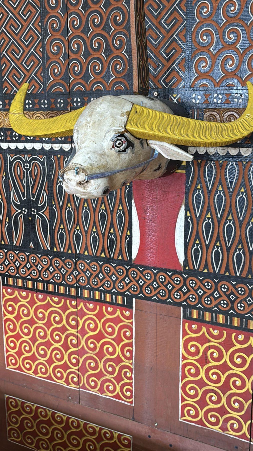 A close-up of a carved wooden buffalo head, its horns painted yellow, mounted on a wall of Toraja geometric patterns.