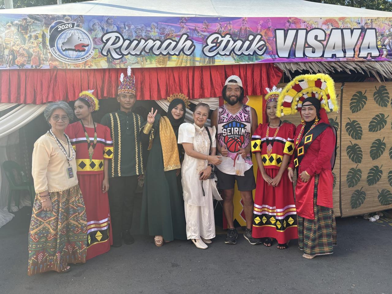 A group photo at the Rumah Etnik Visaya booth, the author standing among hosts and performers dressed in red, black, and gold traditional Visayan costumes.