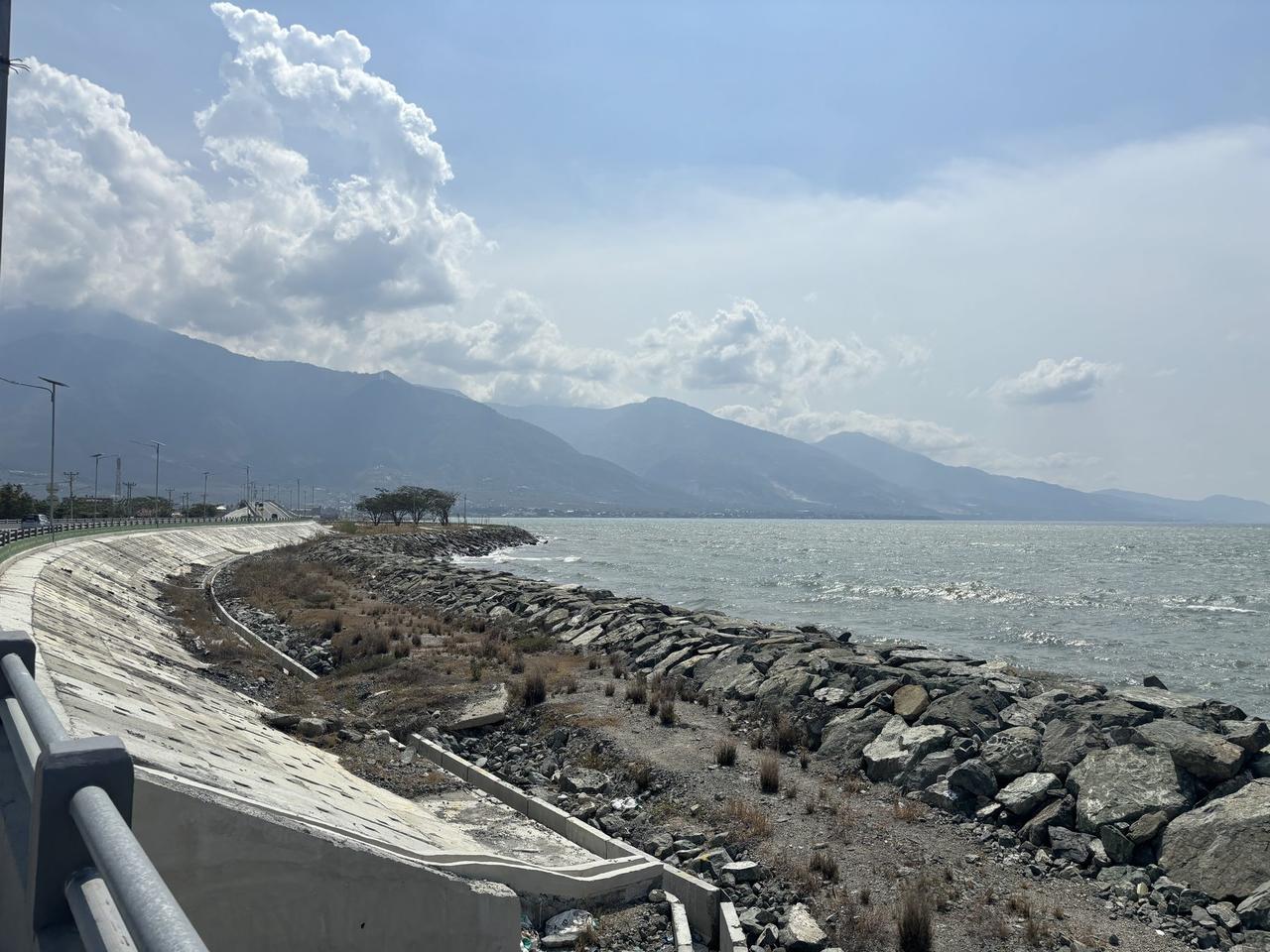 Tanggul laut dan jalan setapak melengkung di sepanjang Teluk Palu, dengan pegunungan menjulang di seberang.