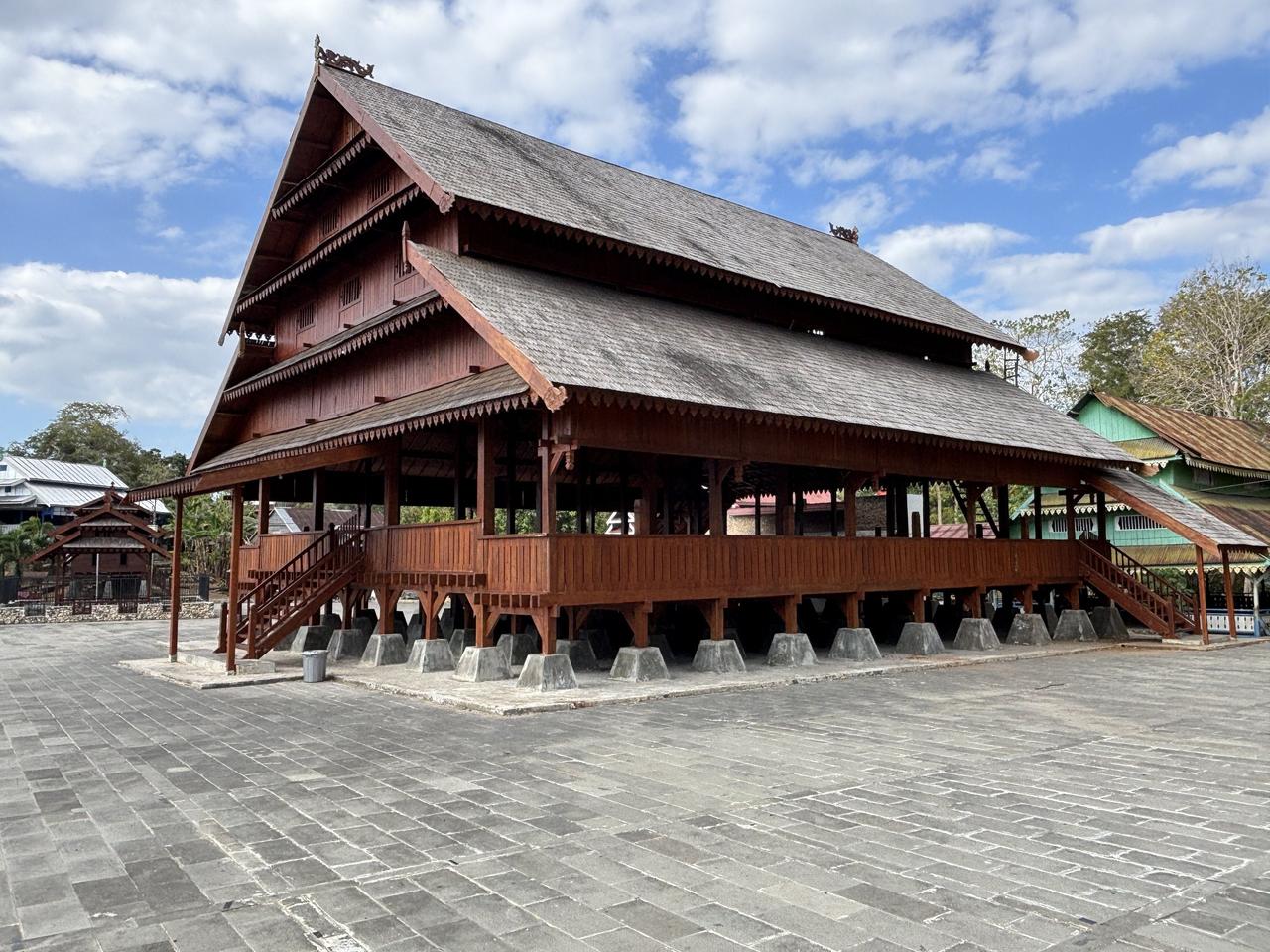 Sebuah balai kayu tradisional yang berdiri di atas tiang panggung, atapnya yang panjang berukir dan bertingkat dalam gaya lokal.