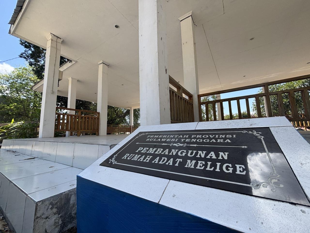 Sebuah prasasti batu bertuliskan Pembangunan Rumah Adat Melige, dengan paviliun istirahat berwarna putih berdiri di samping area istana.