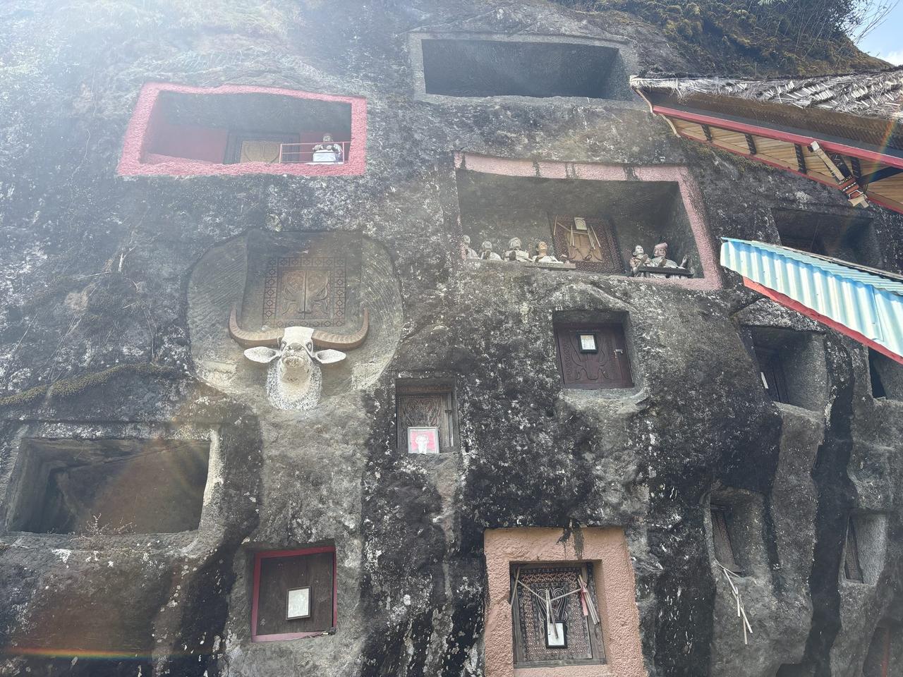 A red-framed chamber holding tau-tau figures beside an ornately carved wooden door set into the rock.