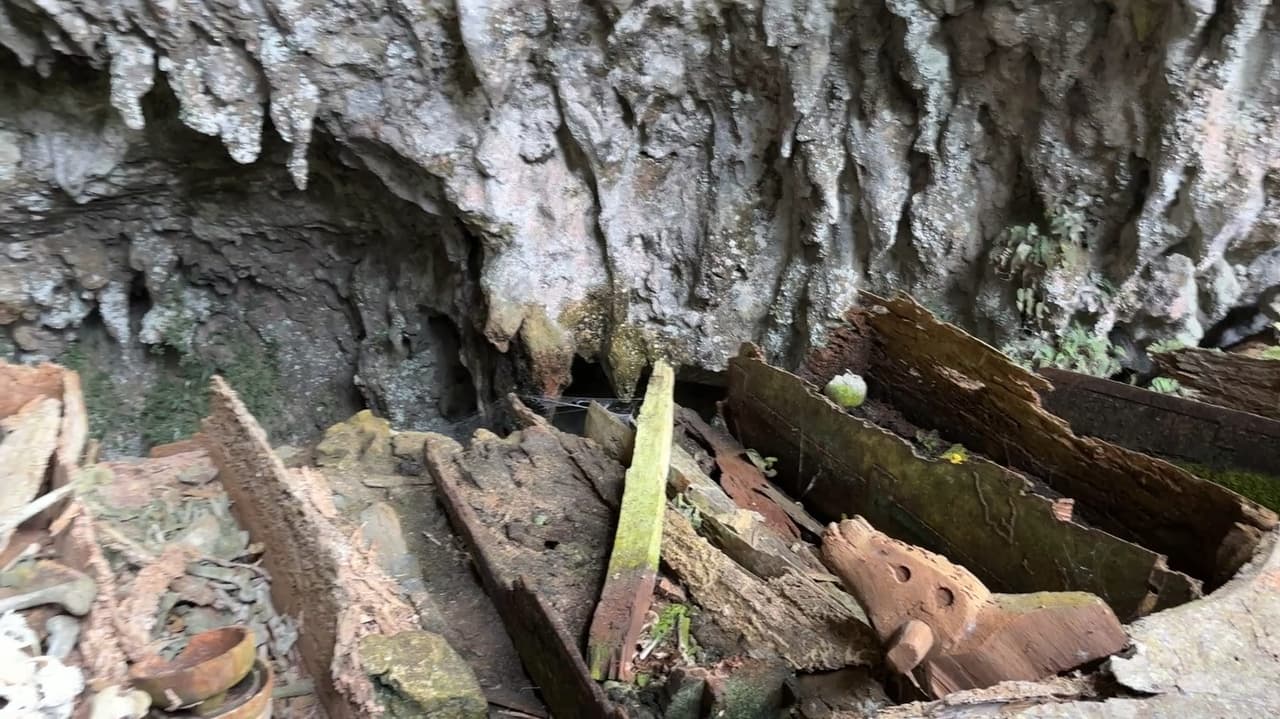 Weathered wooden coffins and carved fragments scattered across the cave floor, stalactites hanging overhead.