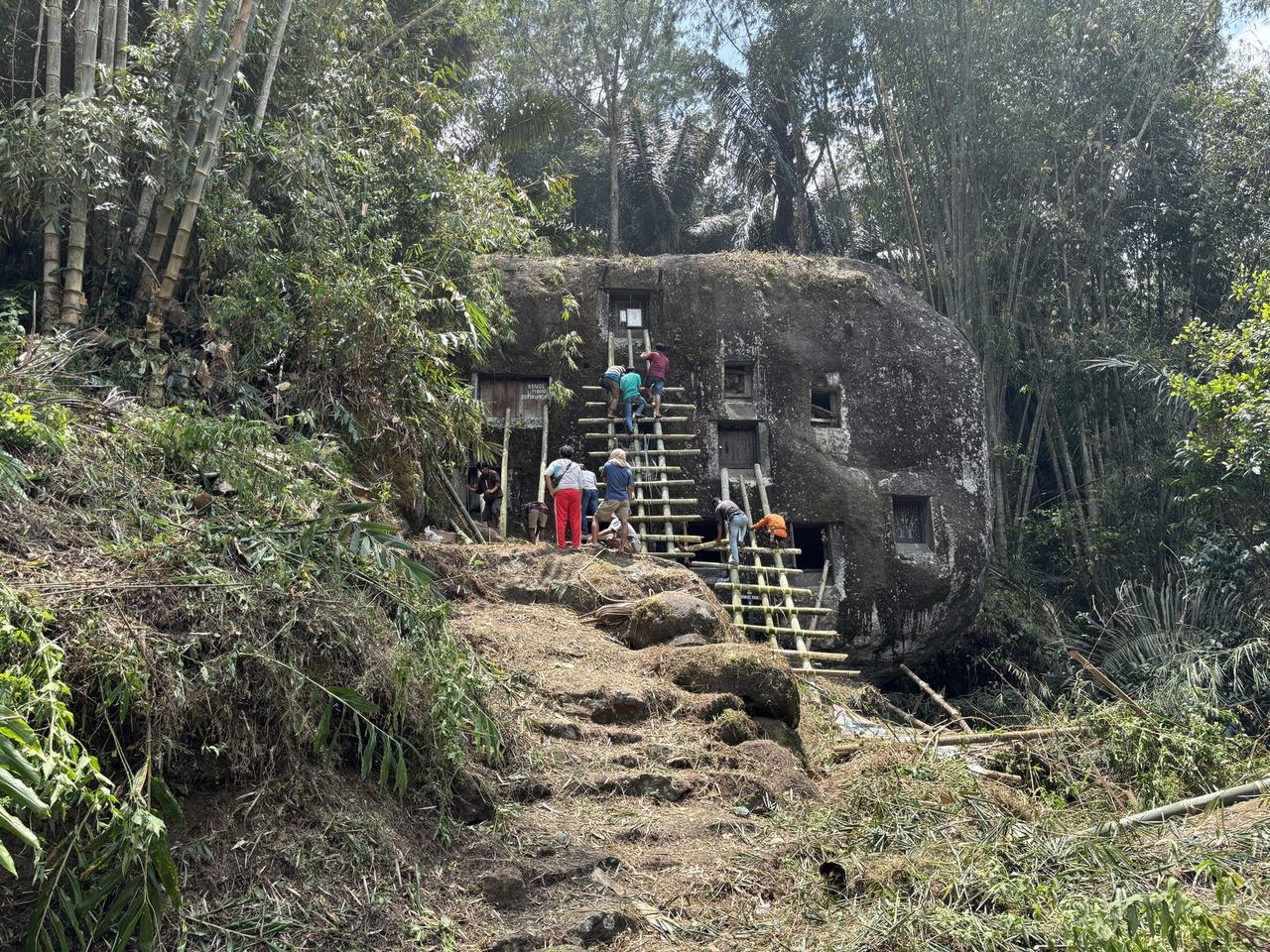 Several bamboo ladders lashed together and leaning against a large mossy boulder riddled with rectangular tomb openings, family members climbing toward different doors.