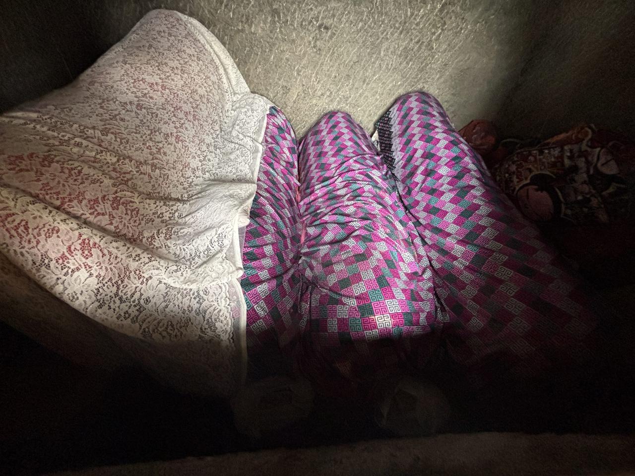 A dim, close view into an opened tomb chamber, coffins wrapped in white lace and pink-and-teal patterned cloth pressed close together.