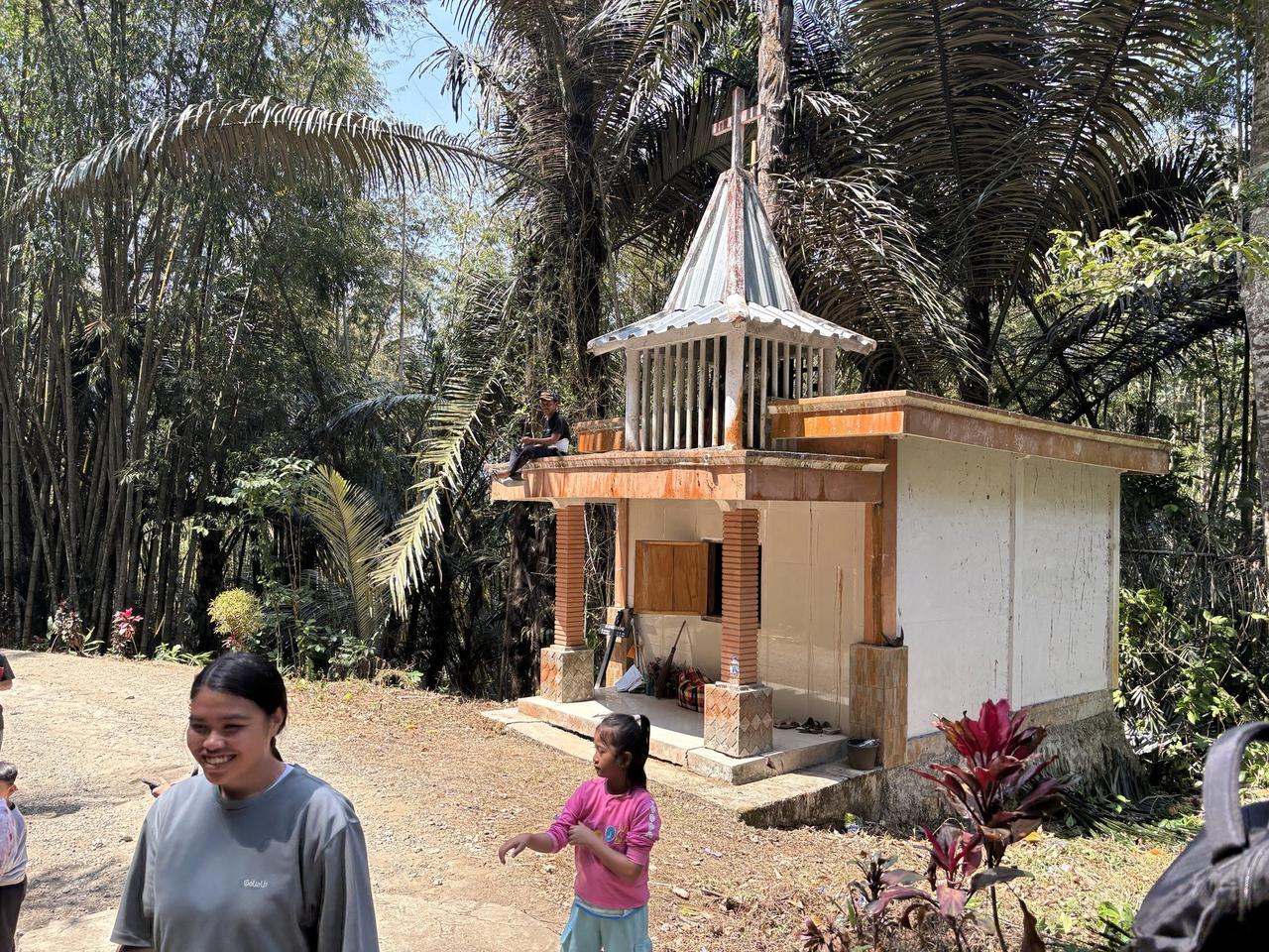 A small white house-shaped Patane tomb with a peaked tin roof and cross, people gathered near its entrance at the base of the hillside site.