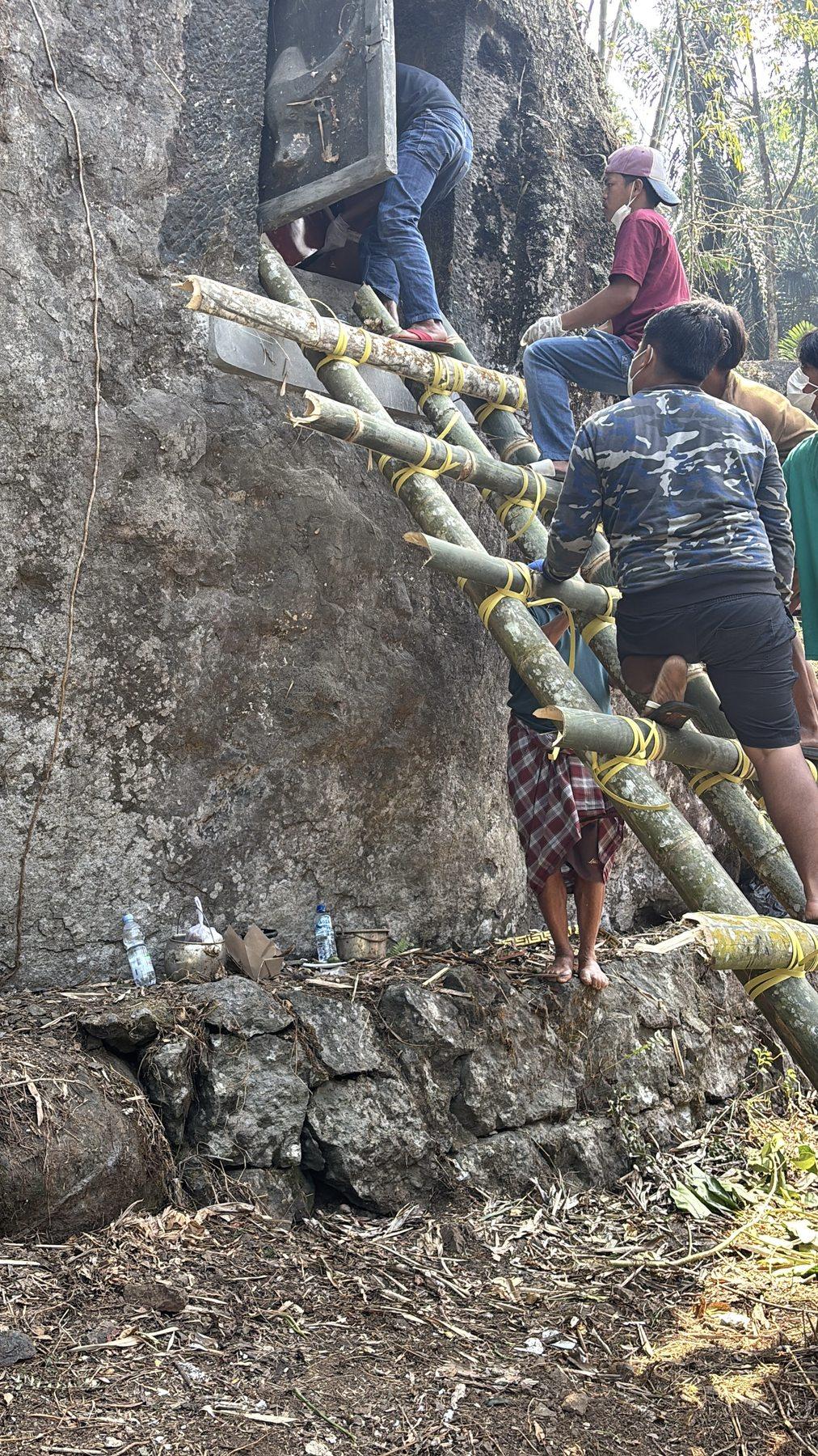 A family member reaching into an opened tomb high on the boulder while others steady the bamboo ladder below.