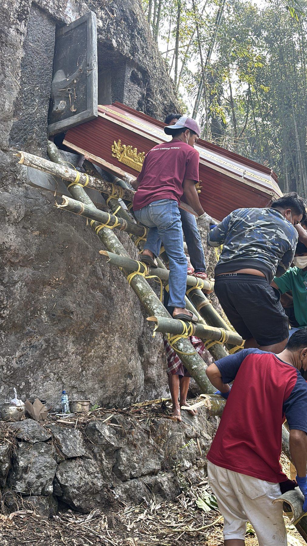 Two men carefully lowering a dark red coffin down a lashed bamboo ladder against the boulder face.