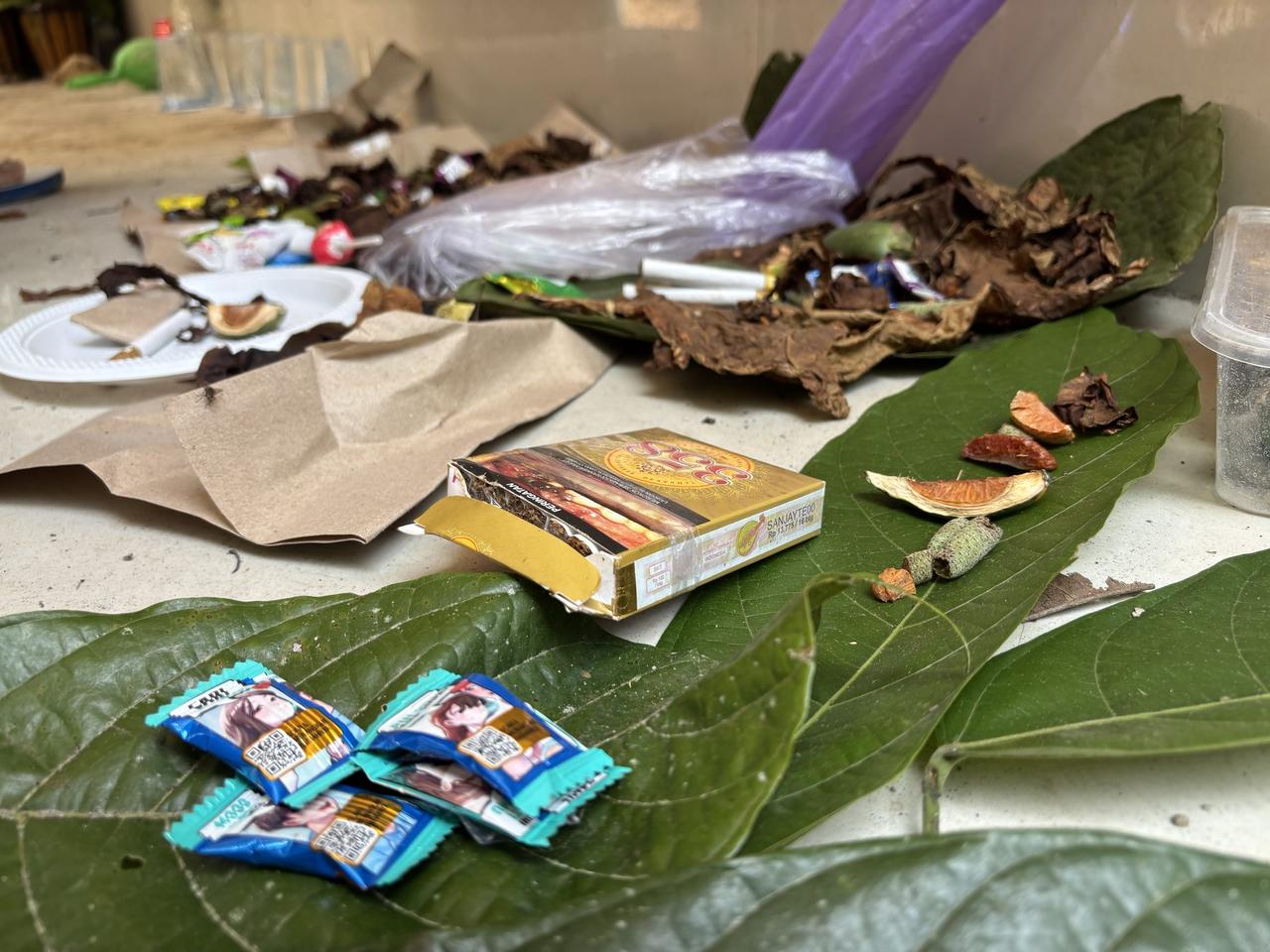 A close view of betel leaf offerings, tobacco, dried fruit peel, and snack wrappers laid out on broad green leaves.