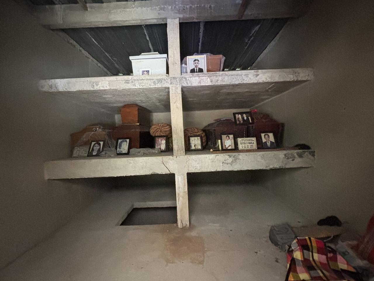 The dim interior of the Patane tomb, concrete shelves holding several coffins and urns each with a small photograph.