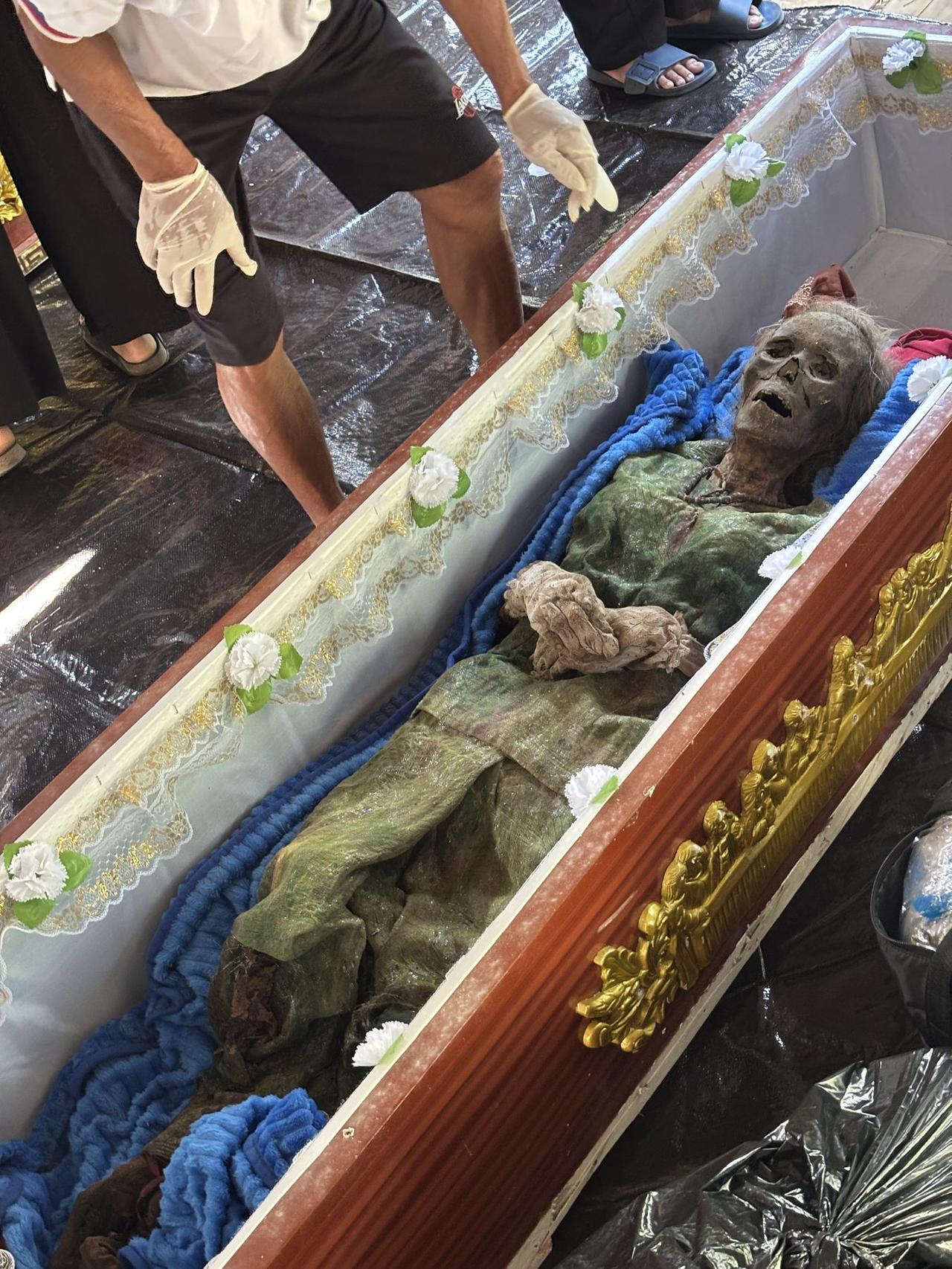 A close view of a mummified figure resting in an open coffin, wrapped in green and blue cloth against white lace trim.