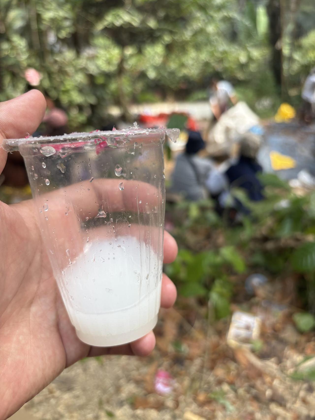 A hand holding a small plastic cup of cloudy white baro palm wine, with people gathered in the background.