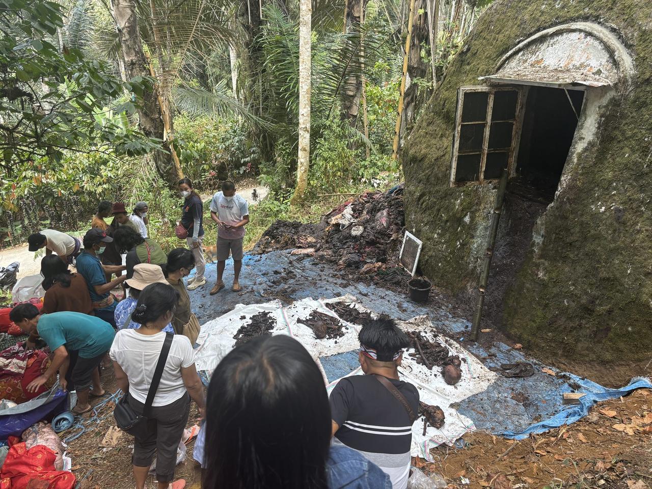 Family members gathered around an open tomb in a mossy boulder, a pile of old debris removed from inside spread on tarps beside them.