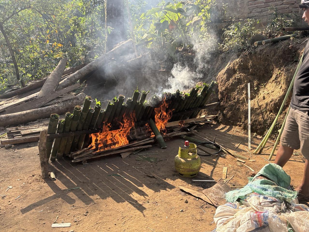 A long row of bamboo tubes propped over an open wood fire, flames and smoke rising as they roast.