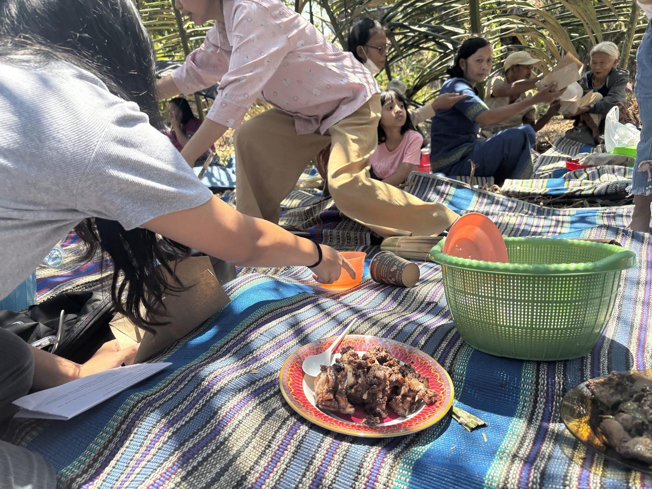 People sitting on woven mats in the shade sharing a meal of buffalo and pork.