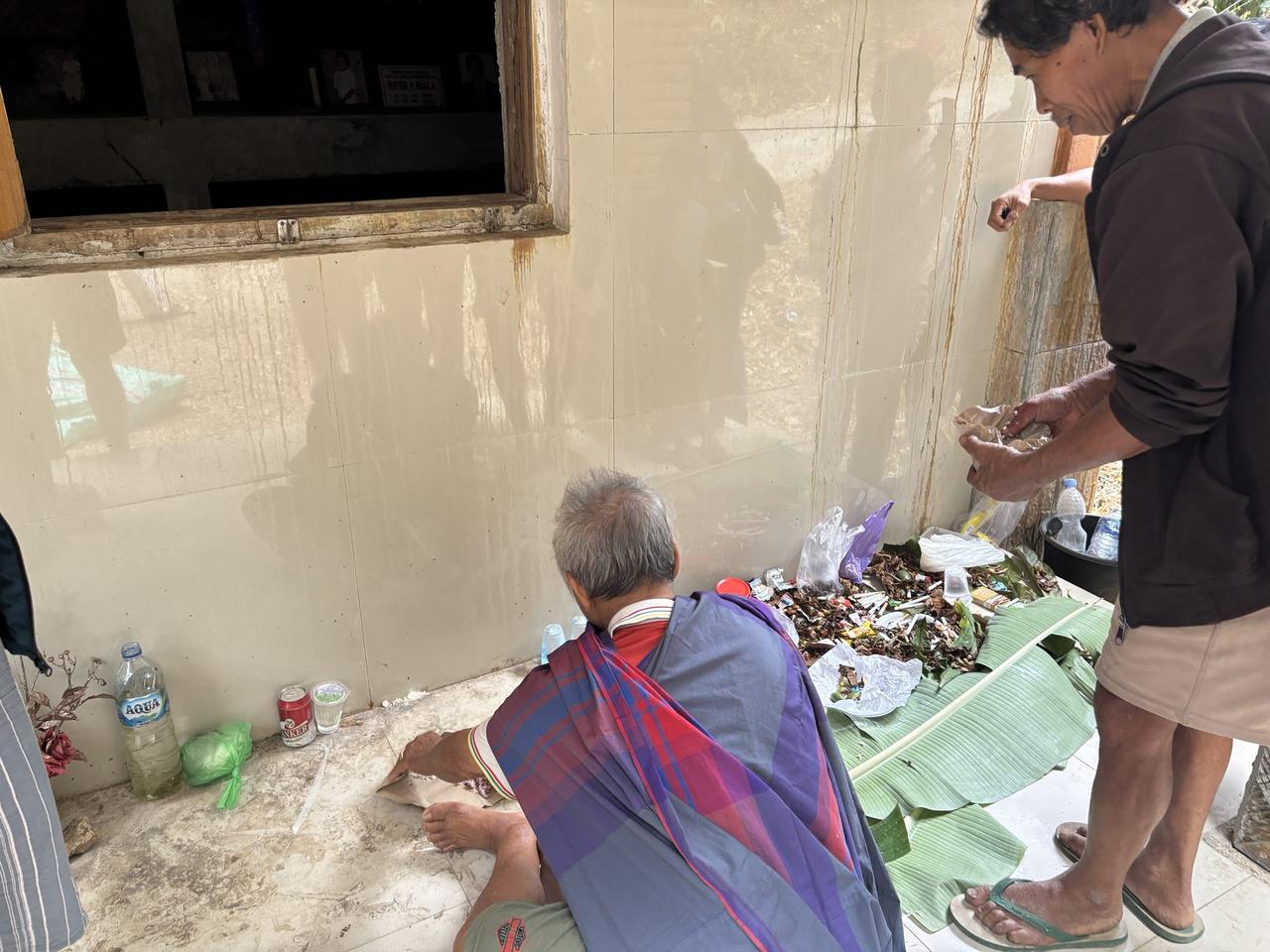 An elderly man seated beside a pile of betel leaf offerings, drink bottles, and wrappers against a tiled wall.
