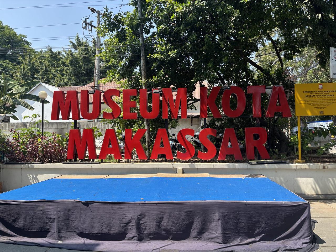 Tulisan berwarna merah pada papan nama Museum Kota Makassar di luar gedung, dikelilingi pepohonan.