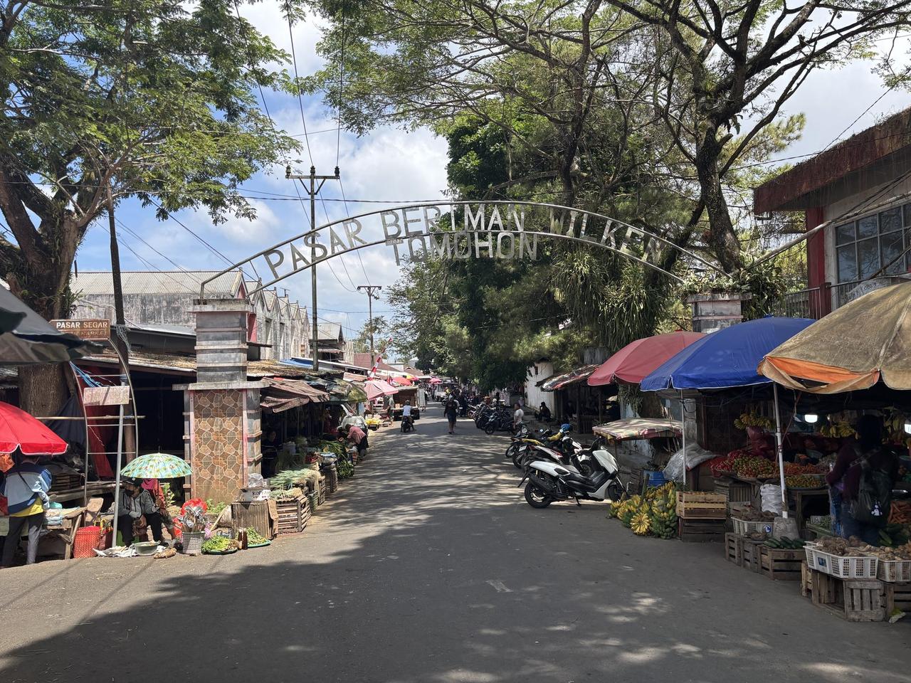 Gapura besi yang menandai pintu masuk Pasar Beriman Wilken Tomohon, dengan lapak-lapak pasar, sepeda motor, dan pisang yang digantung memadati kedua sisi jalan di bawahnya.