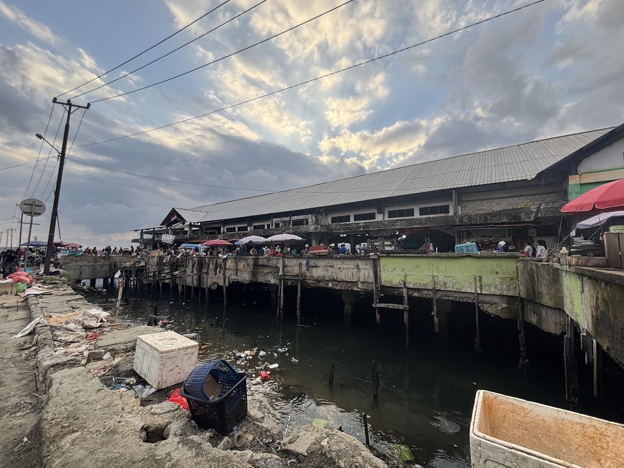 A market building on stilts over the water at Pasar Mardika in Ambon at dusk, trash visible floating in the water below.