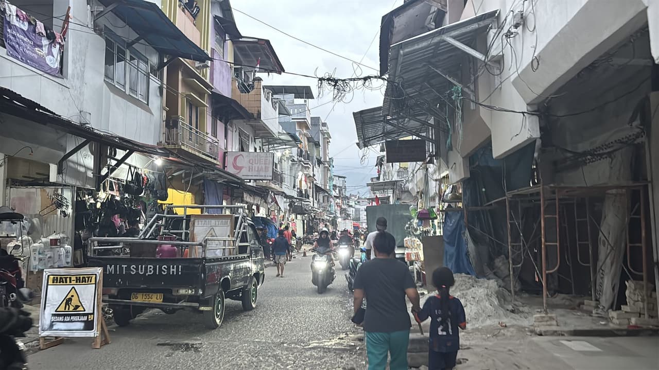 A narrow shopping street inside Pasar Mardika in Ambon, lined with shops, motorbikes, and a delivery truck, a parent and child walking through.