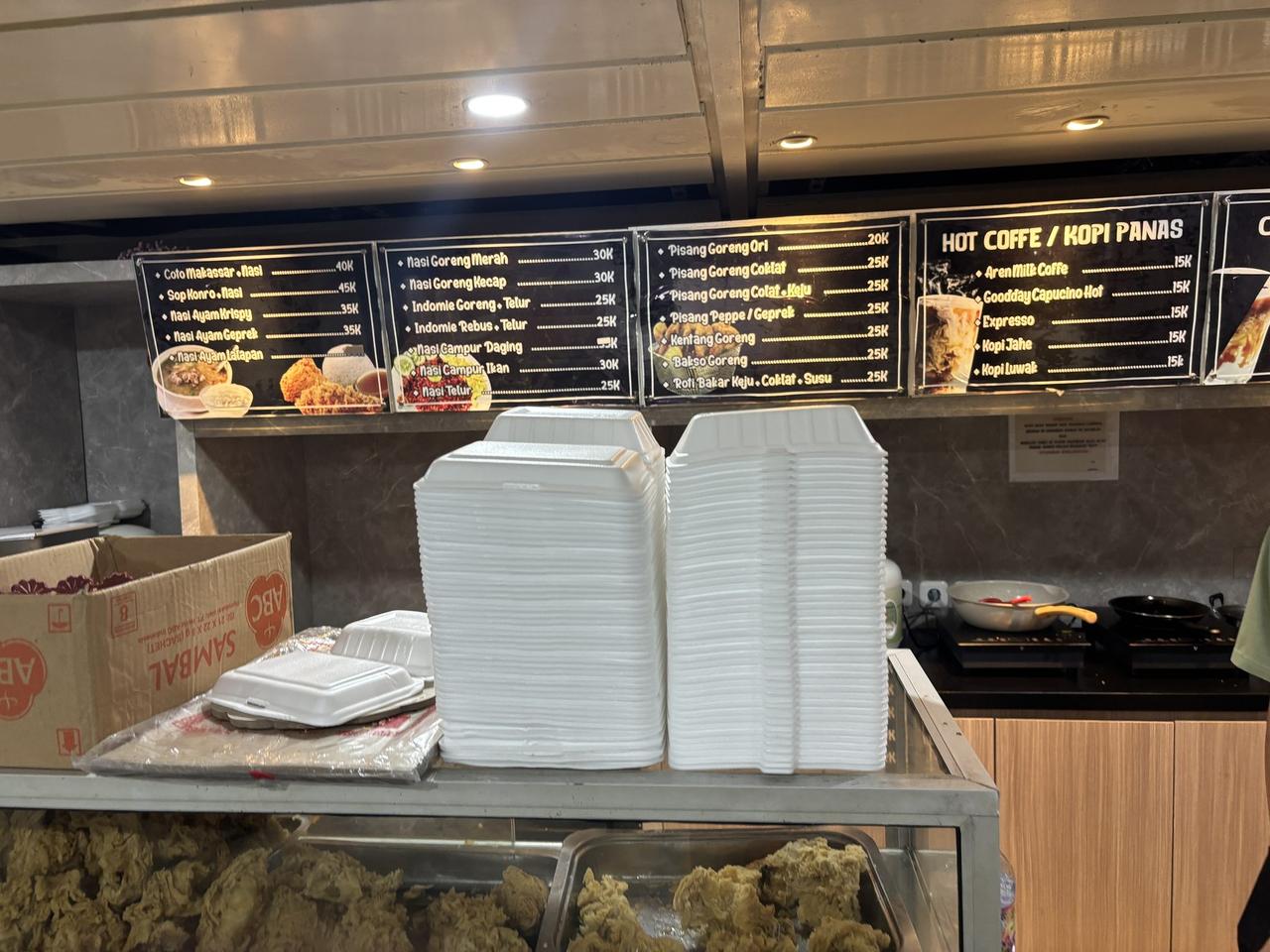 A food stall menu board aboard a PELNI ship listing Coto Makassar, Sop Konro, fried rice, banana fritters, and hot coffee, with stacked styrofoam containers below.