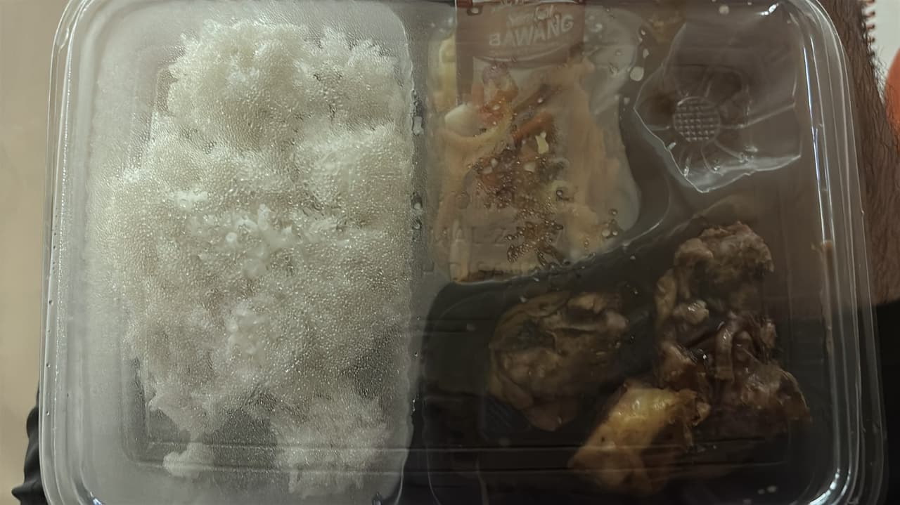 A close-up of a meal tray with rice, a small packet of fried garlic sambal, and pieces of meat, condensation fogging the plastic lid.