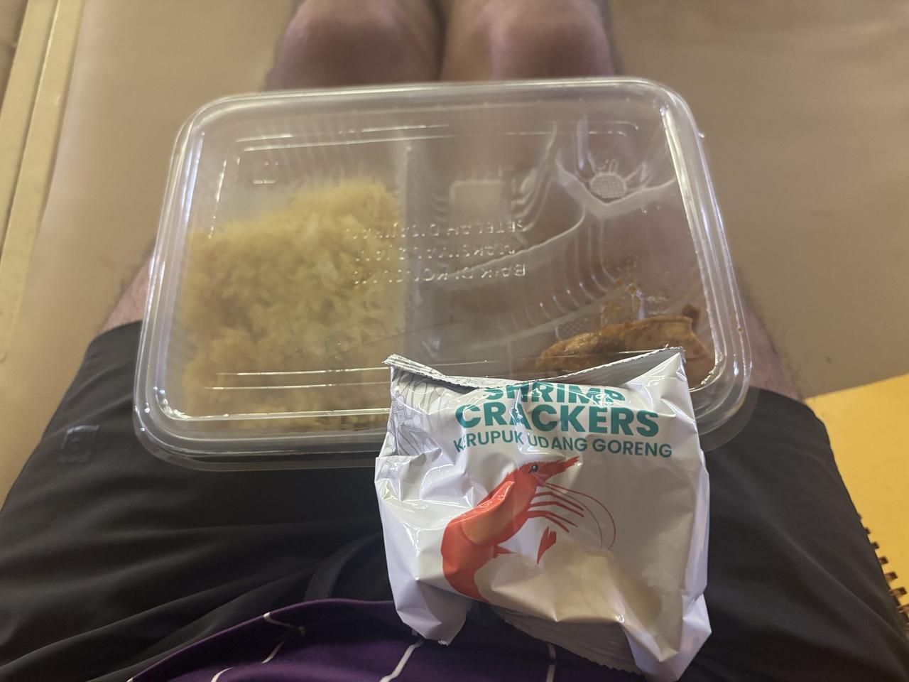 A meal tray of rice and fried chicken on the author's lap, next to an open bag of shrimp crackers.