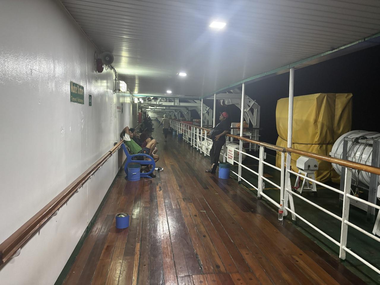 A dim, narrow deck aboard a PELNI ship at night, passengers resting against the wall near covered life rafts.