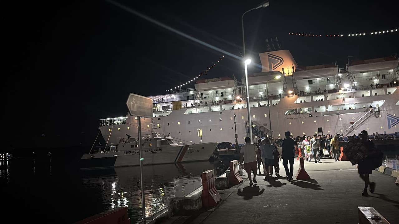A PELNI ship lit up at night at the dock, passengers walking along the pier toward it carrying luggage.