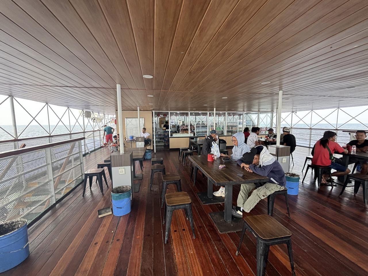 An outdoor deck canteen aboard a PELNI ship, passengers seated at wooden tables and metal stools with the open ocean visible through the railing.