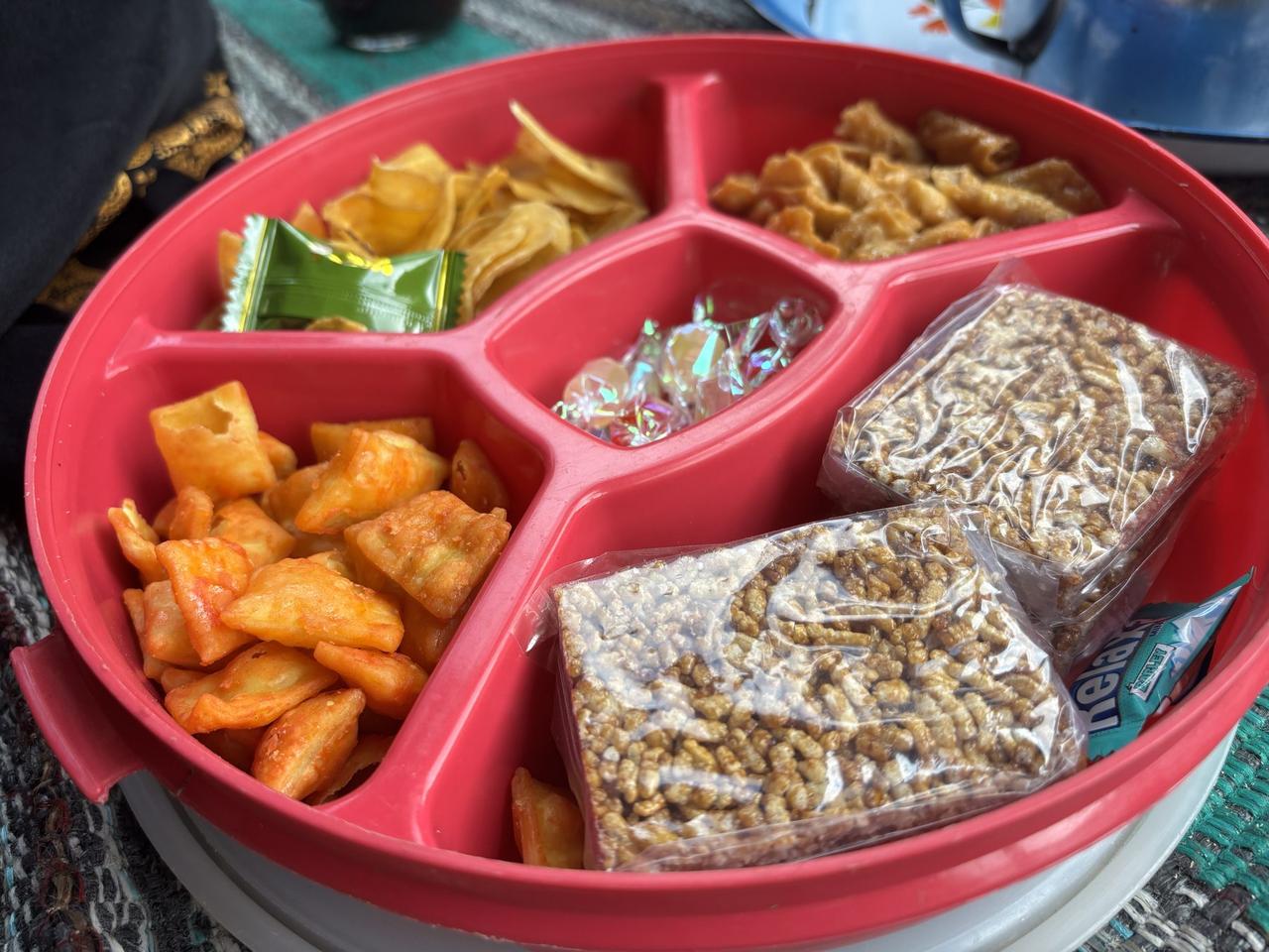 A red plastic tray divided into sections holding local snacks, crackers, and wrapped candies, set out for arriving guests.